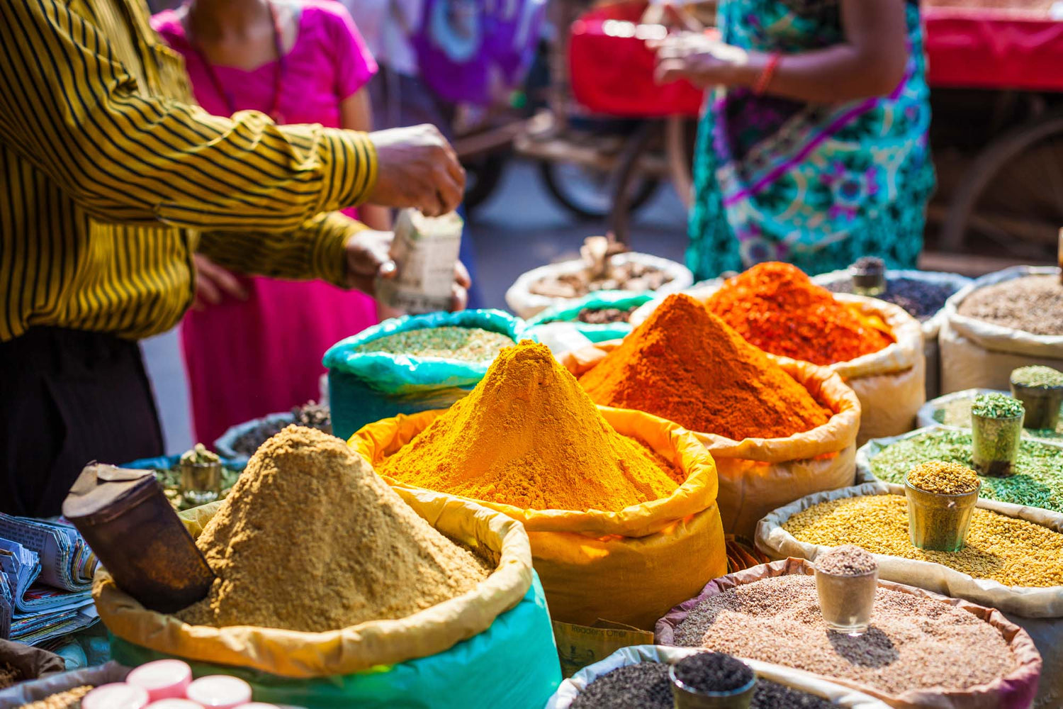 Colorful spices in bags at a market stall with people in the background.
